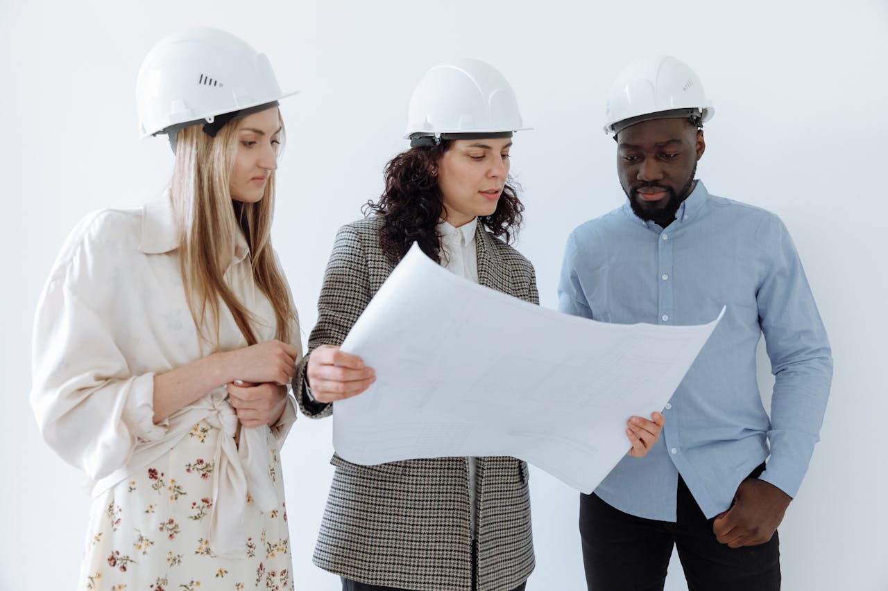 hero-img-02 Three architects wearing hard hats reviewing blueprints indoors for a construction project.